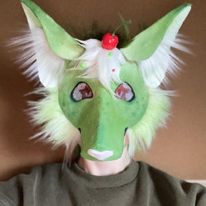 Puede incluir: Una m&aacute;scara peluda verde y blanca con orejas grandes, dise&ntilde;ada para parecerse a una criatura de fantas&iacute;a. La m&aacute;scara presenta una cereza en la parte superior y est&aacute; decorada con detalles verdes y rojos. La m&aacute;scara la lleva una persona con una camisa verde.