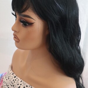 May include: A black wig with long, wavy hair and bangs. The wig is styled on a mannequin head with a white and black polka dot scarf.