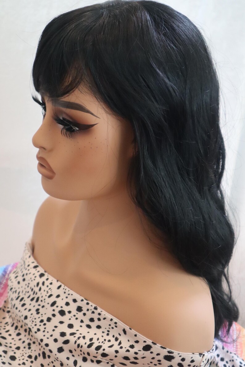 May include: A black wig with long, wavy hair and bangs. The wig is styled on a mannequin head with a white and black polka dot scarf.