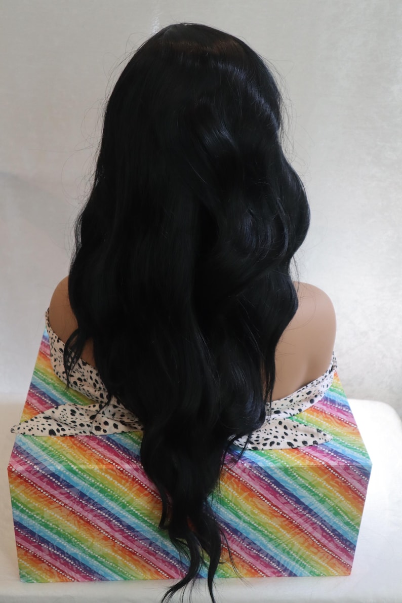 May include: A long, black wig with loose waves. The wig is styled in a way that creates volume and movement. The wig is displayed on a mannequin head with a colorful background.