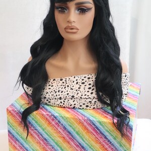 May include: A black wig with long, wavy hair and a side-swept fringe. The wig is displayed on a mannequin head with a white and black patterned top. The mannequin head is sitting on a box with a rainbow striped pattern.