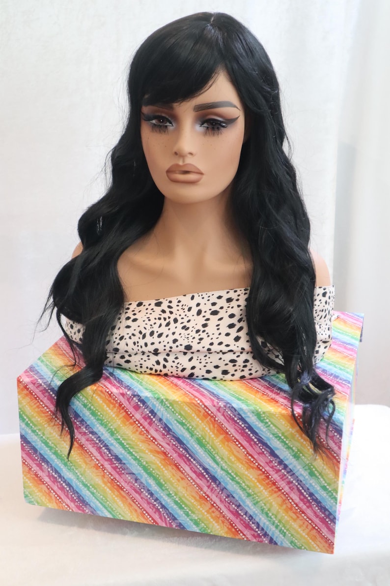 May include: A black wig with long, wavy hair and a side-swept fringe. The wig is displayed on a mannequin head with a white and black patterned top. The mannequin head is sitting on a box with a rainbow striped pattern.