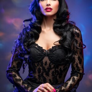 May include: A woman with long black hair and dark red lipstick wears a black lace dress with a low neckline. The dress has long sleeves and a ruffled skirt.