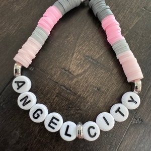 May include: A pink, gray, and white beaded bracelet with silver clasps. The beads spell out "ANGEL CITY".
