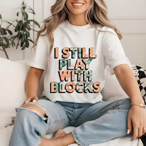 May include: A white t-shirt with the text "I still play with blocks" printed in colorful letters. The letters are decorated with a geometric pattern.