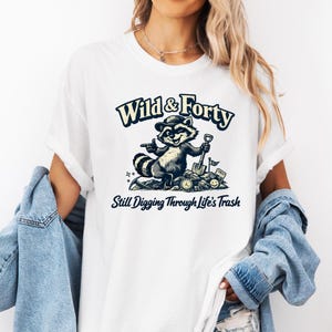 May include: White t-shirt with a raccoon graphic wearing a hat and holding a shovel. The text "Wild & Forty" and "Still Digging Through Life's Trash" are printed above and below the raccoon illustration.