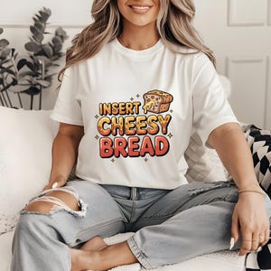 May include: White t-shirt with the text "INSERT CHEESY BREAD" in orange and red, with a cartoon image of cheesy bread. The shirt is worn with light blue jeans.