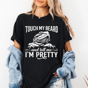 May include: Black t-shirt with the text "TOUCH MY BEARD and tell me I'M PRETTY" in white. A detailed illustration of a bearded dragon is centered below the text. The shirt is a casual, unisex style.