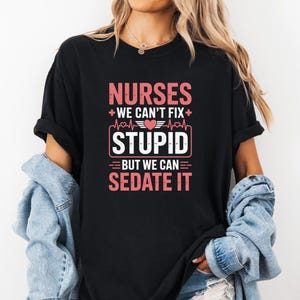 以下が含まれることがあります： ピンクと白で「NURSES we can't fix STUPID but we can sedate it」というテキストがプリントされた黒いTシャツ。デザインには、心臓、心拍ライン、十字のシンボルが含まれています。Tシャツは人が着ています。