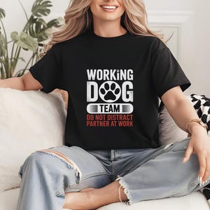 May include: Black t-shirt with the text "WORKING DOG TEAM" in white, featuring a paw print graphic. Below the text is "DO NOT DISTRACT PARTNER AT WORK" in red. The shirt is worn by a person.