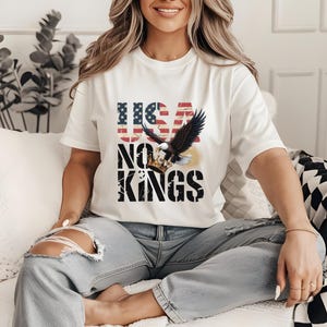 May include: White t-shirt featuring a graphic design with the text "USA NO KINGS". The design includes an American flag and an eagle with a crown. The shirt is worn by a person wearing ripped blue jeans.