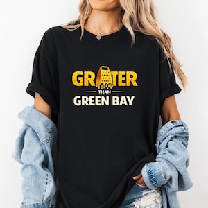 May include: Black t-shirt with the text "GRATER than GREEN BAY" in yellow and white. The word "GRATER" features a yellow cheese grater graphic. The shirt is worn by a person with blonde hair and a denim jacket.