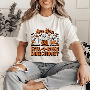 May include: White t-shirt with a Halloween-themed graphic. The design features three cartoon ghosts, a pumpkin, and the text "Are You Fall-O-Ween Directions?" in orange and black. The ghosts are holding coffee, a pumpkin, and a book.