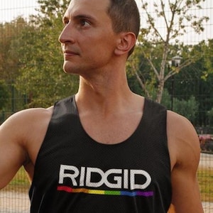 May include: Black mesh tank top with the word "RIDGID" in white block letters and a rainbow stripe underneath. The tank top is worn by a person outdoors near a basketball court.