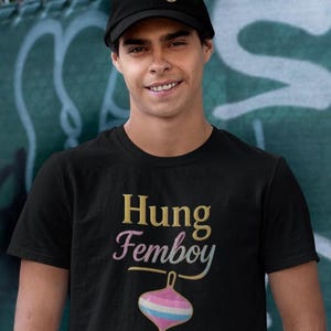 May include: A black t-shirt and baseball cap with the word "Hung" in gold lettering. The t-shirt features the words "Hung Femboy" and a colorful spinning top graphic.