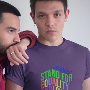 May include: A purple t-shirt with the text "ALWAYS STAND FOR EQUALITY AND LOVE NEVER HATE" in a colorful, 3D design. The shirt is worn by a person, with another person's arm resting on their shoulder.