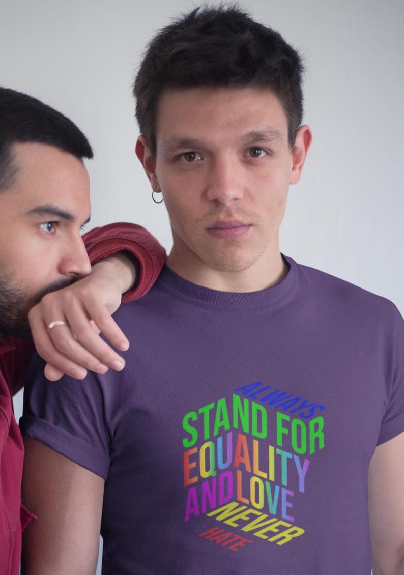 May include: A purple t-shirt with the text "ALWAYS STAND FOR EQUALITY AND LOVE NEVER HATE" in a colorful, 3D design. The shirt is worn by a person, with another person's arm resting on their shoulder.