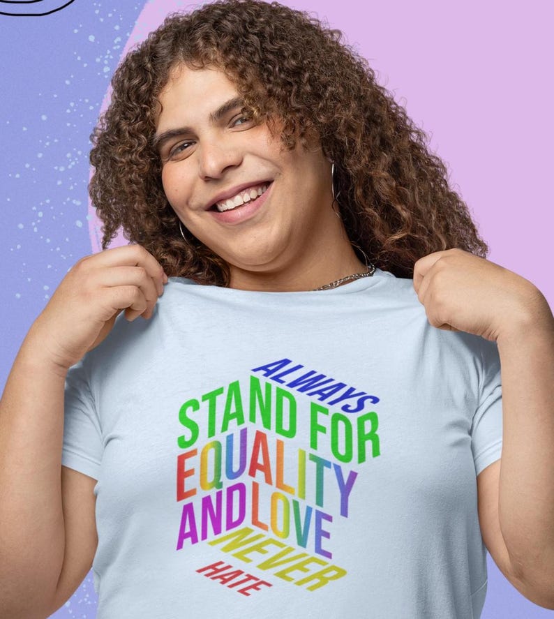 May include: Light blue t-shirt with the text "ALWAYS STAND FOR EQUALITY AND LOVE NEVER HATE" in a rainbow gradient. The shirt is being held up by a person with curly brown hair.