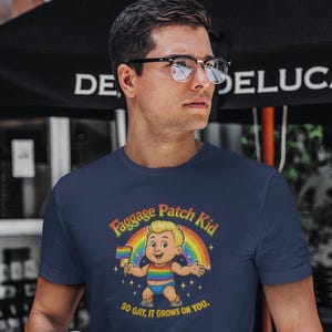 May include: Navy blue t-shirt with a cartoon design of a child holding a rainbow flag, with the text "Faggage Patch Kid" above and "SO GAY, IT GROWS ON YOU." below. The design includes a rainbow and stars.