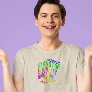 May include: A light beige t-shirt with the text "ALWAYS STAND FOR EQUALITY AND LOVE NEVER HATE" in a rainbow color scheme. The shirt is being worn by a person with dark hair and fair skin.