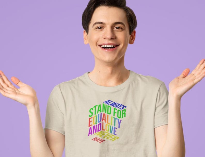 May include: A light beige t-shirt with the text "ALWAYS STAND FOR EQUALITY AND LOVE NEVER HATE" in a rainbow color scheme. The shirt is being worn by a person with dark hair and fair skin.
