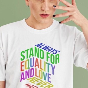 May include: White t-shirt with the text "ALWAYS STAND FOR EQUALITY AND LOVE NEVER HATE" in a rainbow gradient. The text is arranged in a 3D block design. The shirt is being worn.
