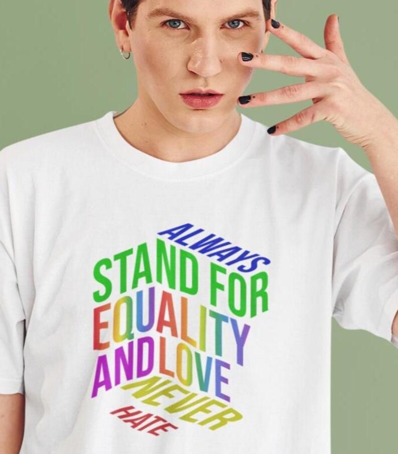 May include: White t-shirt with the text "ALWAYS STAND FOR EQUALITY AND LOVE NEVER HATE" in a rainbow gradient. The text is arranged in a 3D block design. The shirt is being worn.