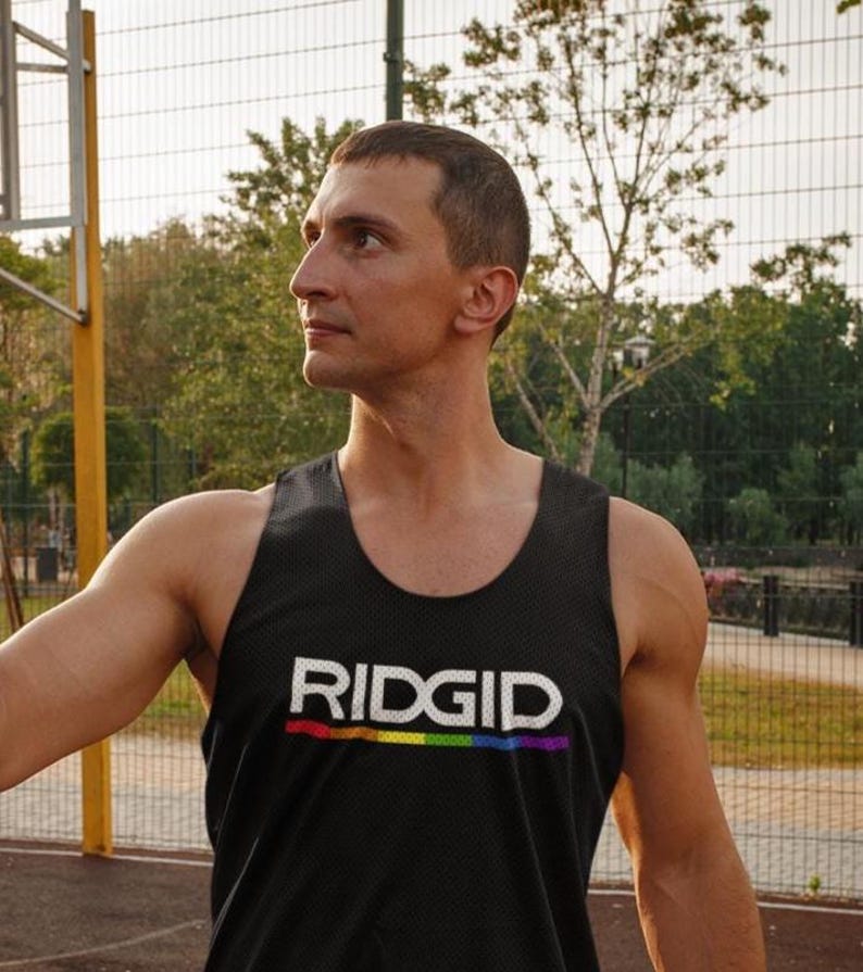 May include: Black mesh tank top with the word "RIDGID" in white block letters and a rainbow stripe underneath. The tank top is worn by a person outdoors near a basketball court.