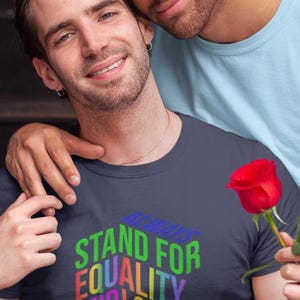 May include: A navy blue t-shirt with the words "STAND FOR EQUALITY AND LOVE NEVER HATE" in rainbow colors. A red rose is held in the hand. Two people are in the image.