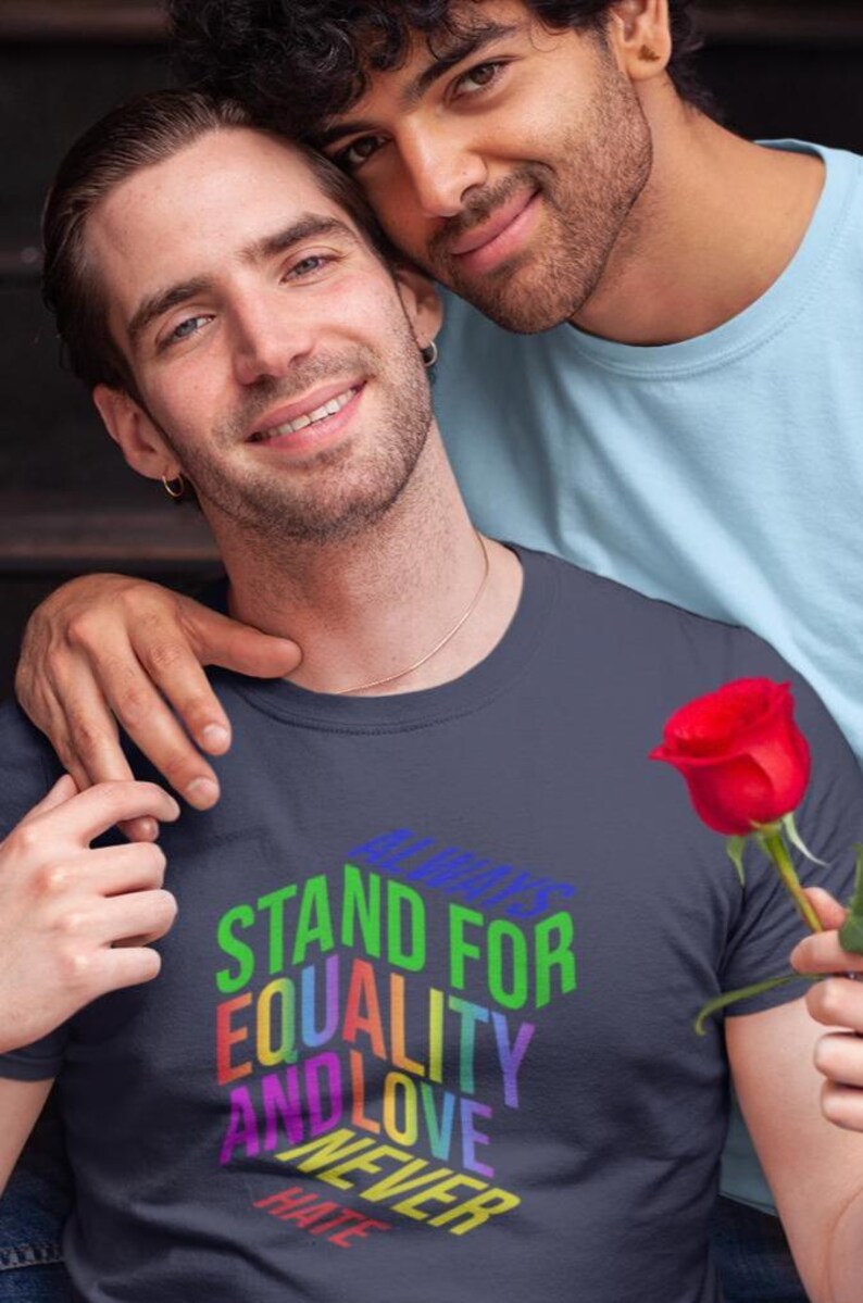 May include: A navy blue t-shirt with the words "STAND FOR EQUALITY AND LOVE NEVER HATE" in rainbow colors. A red rose is held in the hand. Two people are in the image.