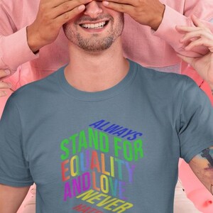 May include: A slate blue t-shirt with the text "ALWAYS STAND FOR EQUALITY AND LOVE NEVER HATE" in a rainbow gradient. The shirt is being worn by a person with a beard, and another person is covering their eyes.