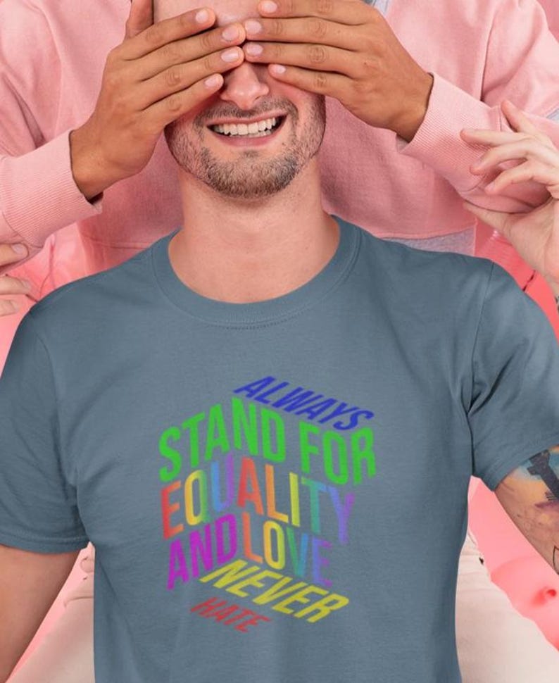 May include: A slate blue t-shirt with the text "ALWAYS STAND FOR EQUALITY AND LOVE NEVER HATE" in a rainbow gradient. The shirt is being worn by a person with a beard, and another person is covering their eyes.