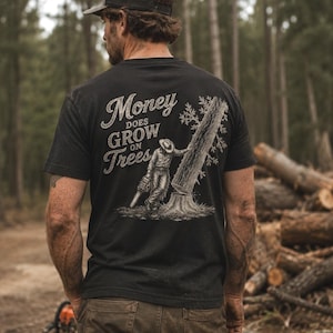 Peut inclure: Un t-shirt noir à manches courtes avec un motif au dos. Le dessin représente une personne portant un chapeau et tenant une tronçonneuse près d'une souche d'arbre, avec le texte "Money DOES GROW ON Trees" en police blanche vieillie.