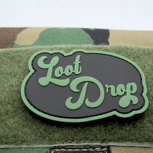 May include: A black and green patch with the text "Loot Drop" in a retro font style. The patch is attached to a green and brown camouflage fabric.