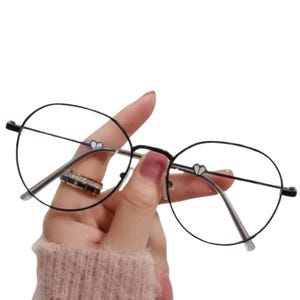 May include: Round, black-rimmed eyeglasses with clear lenses. The frames have small heart-shaped details on the temples. The glasses are held by a hand wearing a ring with black and silver stones. The background is white.