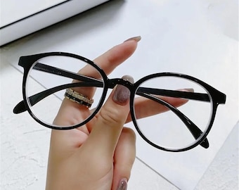 May include: Black-framed eyeglasses with a round lens shape. The glasses are held up by a hand. The frames are thick and have a glossy finish. The lenses are clear. The overall design is modern and stylish.