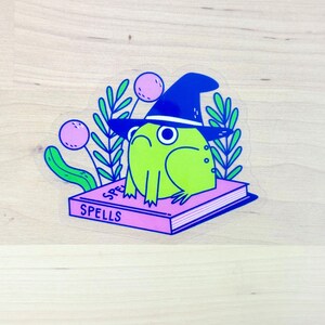 May include: A whimsical sticker featuring a green frog wearing a blue witch hat, perched atop a pink book labeled "SPELLS." The frog is surrounded by green leaves and pink orbs, creating a magical theme.