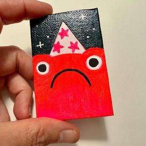 May include: A small, rectangular painting featuring a sad-faced creature. The creature is red with black eyes and a curved mouth. Above it is a dark blue background with a white party hat with pink stars. The painting is held by a hand.