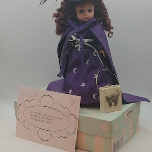 Könnte beinhalten: Eine Madame Alexander Starlight Witch Puppe, die einen schwarzen Hexenhut, einen lila Umhang mit Mond- und Sternakzenten trägt und eine kleine Handtasche hält. Die Puppe befindet sich auf einer Schachtel mit Echtheitszertifikat.