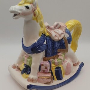 Könnte beinhalten: Keramik-Schaukelpferd-Figur mit weißem Körper, gelber Mähne und Schweif sowie einer blauen Satteldecke. Das Pferd ist mit rosa, grünen und goldfarbenen Geschenken verziert. Der Sockel ist weiß mit blauem Rand.