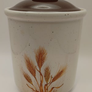 Puede incluir: Un tarro de cerámica blanco con tapa marrón y diseño de trigo. El tarro tiene un esmalte moteado y mide aproximadamente 18 cm de alto.