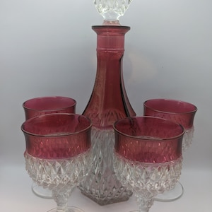 May include: A vintage glass decanter set in ruby red. The set includes a diamond-cut decanter with a faceted stopper, and four matching wine glasses. The glasses and decanter have a clear base and ruby red rims. A classic glassware set.
