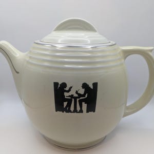Puede incluir: Una tetera de cerámica blanca con un borde plateado alrededor de la tapa y la base. La tetera presenta una silueta negra de dos personas sentadas a una mesa.