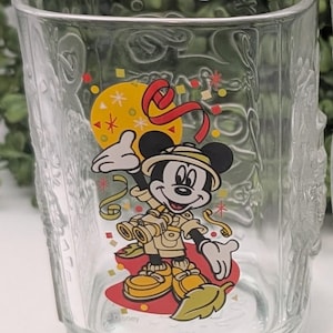 Puede incluir: Un vaso de vidrio transparente con un diseño de Mickey Mouse. Mickey está vestido de explorador y sostiene unos prismáticos. El vaso tiene confeti y una cinta roja. La parte inferior del vaso dice "Disney" y "2000".