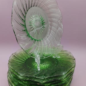 Può includere: Un set di otto piatti in vetro verde con un piatto in vetro trasparente in cima. I piatti hanno un design a spirale e un motivo a stella al centro.