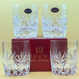 Op de afbeelding: Set van vier heldere kristallen glazen met een gesneden ontwerp. De glazen staan op een rode doos met de tekst "LENOX BAR CRYSTAL" op de voorkant.