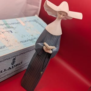 Puede incluir: Figura de porcelana de una monja con hábito gris y toca blanca, sosteniendo un libro y un rosario. La figura tiene una expresión serena y está sobre un fondo rojo. La figura mide aproximadamente 25 cm de altura.