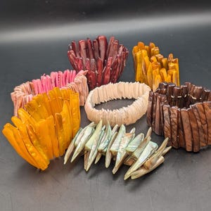 Puede incluir: Una colección de pulseras artesanales en varios colores, incluyendo rosa, naranja, marrón y verde iridiscente. Las pulseras están hechas de materiales naturales, posiblemente conchas o madera, y tienen una apariencia texturizada y orgánica. Se muestran sobre una superficie oscura.
