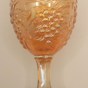 Op de afbeelding: Een vintage, iriserende, perzikkleurige glazen kelk met een druivenranken- en druivenontwerp. De kelk heeft een lange, slanke steel en een brede, ronde voet.