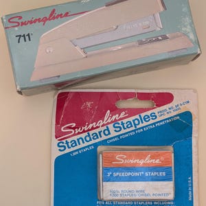May include: A vintage Swingline 711 stapler in its original box, alongside a package of Swingline Standard Staples. The stapler is beige and silver, with the brand name prominently displayed. The staple package features red, blue, and white packaging.